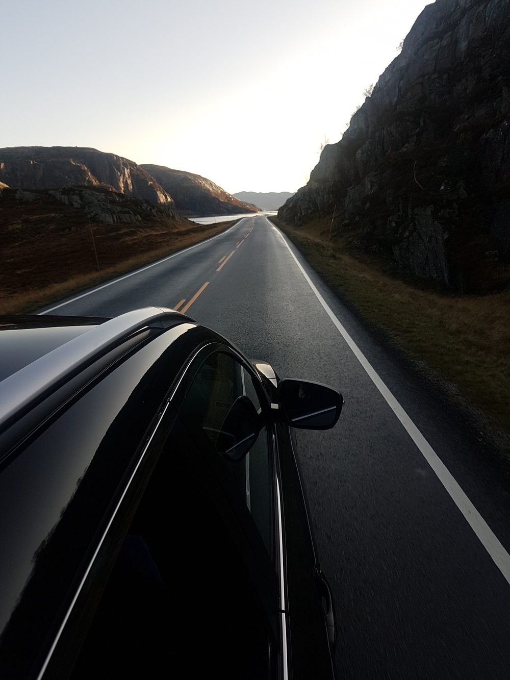 Utsikt fra bilvindu langs norsk kystvei ved solnedgang, med fjell og fjord